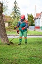 Man in overall and safety helmet trims overgrown lawn by grass cutter