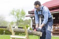 Man in overalls saws a tree on sawhorses in courtyard with a chainsaw.