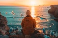 A man in an oversized hoodie sits on the edge of a cliff watching seagulls flying over the sea at sunset Royalty Free Stock Photo