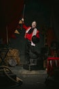 Man with pale face makeup, in red stage costume and hat making creative performance over dark retro circus backstage Royalty Free Stock Photo