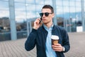 A man with a paper Cup of coffee, goes through the city, a handsome guy in stylish clothes and sunglasses, making a phone call Royalty Free Stock Photo