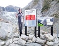 A Man photo stand shows warning sign regulation for ice fall, Rock fall, flooding, river surge at Fox Glacier.