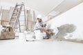 Man plasterer construction worker at work, takes plaster from bucket and puts it on trowel to plaster the wall, wears helmet