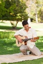 Mid adult man kneeling on picnic blanket in park, strumming guitar holding red rose between teeth Royalty Free Stock Photo