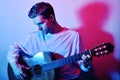 A man plays an acoustic guitar in neon red-blue light. A man learns to play the guitar, music, hobbies