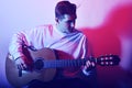 A man plays an acoustic guitar in neon red-blue light. A man learns to play the guitar, music, hobbies