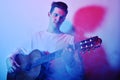 A man plays an acoustic guitar in neon red-blue light. A man learns to play the guitar, music, hobbies