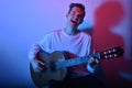 A man plays an acoustic guitar in neon red-blue light. A man learns to play the guitar, music, hobbies