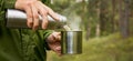 man pours hot tea from thermos flask into a mug in forest. nature tourism and camping concept. copy space