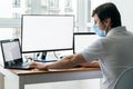 A man in protective blue medical mask is sitting at a workplace with two laptops and a monitor near the window. Office work