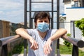 Man in protective blue medical mask standing on the outdoor sports ground, resting his hands on the bars. Male in a respirator