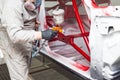 A man in protective overalls and a mask holds a spray bottle in his hand and sprays red paint onto the frame of the car body after
