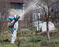 A man in a protective suit and a mask with a pollinator on his back sprays trees and bushes with a chemical solution Royalty Free Stock Photo