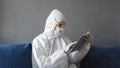 Man in protective suit, medical mask and rubber gloves sits at home and works with tablet on a sofa during quarantine