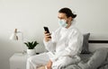 Man in protective white suit and medical mask is using a phone at his home sitting on a bed because of coronavirus Royalty Free Stock Photo