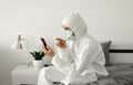 Man in protective white suit and medical mask is using a phone at his home sitting on a bed because of coronavirus Royalty Free Stock Photo