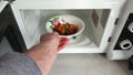 A man puts his hand a white bowl with a pattern with food in the microwave front view, open microwave