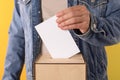 Man putting his vote into ballot box on yellow background, closeup