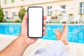 Man relaxing by the outdoor swimming pool with mockup smartphone in hands with blank white touchscreen during summer vacation