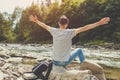 Man relaxing by mountain river enjoying natural landscape. Traveller backpacker raising arms feeling happy. Summer trip