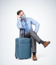 Man in a respiratory mask, shirt and tie sits with a suitcase waiting for a flight. Pandemic Travel concept.