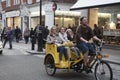 A man riding a Rickshaw with happy women in the back down a street in London