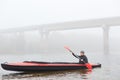 Man rowing in boat on foggy morning, holding paddle in hands, posing in water with bridge on background, looking in front of him, Royalty Free Stock Photo