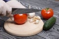 A man in rubber gloves cuts a tomato with a knife on a cutting board. Nearby are cucumber and garlic. On the surface of brushed