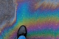 A man's foot steps on a rainbow stain of gasoline on the asphalt.