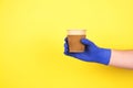 Man`s hand in blue disposable protective gloves on yellow background holding cardboard cup for coffee or other hot drinks take Royalty Free Stock Photo