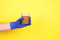 Man`s hand in blue disposable protective gloves on yellow background holding cardboard cup for coffee or other hot Royalty Free Stock Photo