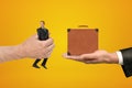 Man`s hand exchanging tiny businessman for tiny brown travel case held in another man`s hand on amber background.