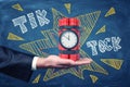 Man`s hand facing up holding dynamite bundle with time bomb on blackboard background with chalk words `tick tock`.