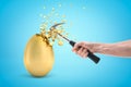 Man`s hand hitting and breaking the top of big gold egg with a hammer on light blue background. Royalty Free Stock Photo