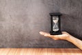 A man`s hand holding a black hourglass on wooden table with old gray concrete walls. Royalty Free Stock Photo