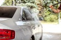 Man`s hand holding hose, spraying water to widow of silver car in self serve carwash, sun lit blurred green trees in background