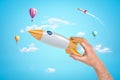 Man`s hand holding silver and yellow space rocket against bright blue sky with hot air balloons and one more rocket