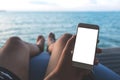 A man`s hand holding white mobile phone with blank desktop screen sitting by the sea and blue sky background Royalty Free Stock Photo