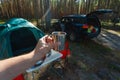 The man's hand holds an iron mug with a warm drink on the background of the table and chairs, campfire, a tent and an SUV in camp Royalty Free Stock Photo