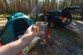 The man's hand holds an iron mug with a warm drink on the background of the table and chairs, campfire, a tent and an SUV in camp Royalty Free Stock Photo