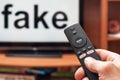 A man`s hand holds a TV remote control, all the buttons on the remote control are signed news, on the TV the inscription is fake
