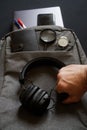 A man`s hand holds wireless headphones on the background of a gray textile backpack with accessories and gadgets for a trip or