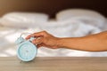 Man`s hand stopping blue classic alarm clock on vintage wooden table on blurred messy white bedding background Royalty Free Stock Photo