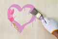 Man`s hand wearing white glove holding old grunge paintbrush and painting pink heart on wooden wall with pink flowers Royalty Free Stock Photo