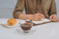 Man`s hand wrote message on white notebook with pen and croissant on wood table, business vintage concept