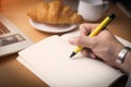 man`s hand wrote message on white notebook with yellow pen and croissant on wood table