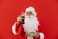 A man in a Santa Claus costume is posing on a red background, holding a bottle of cola and plate of chocolate cookies, offering