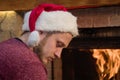 A man in a Santa hat bows his head and looks sad by the burning fireplace. The feast of Christmas alone. Royalty Free Stock Photo