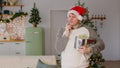 Man in santa hat with gift box standing near Christmas tree in living room. Thoughtful male looks around, scratches chin