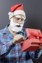 Santa with a white foam beard looks surprised and takes something out of a red gift box